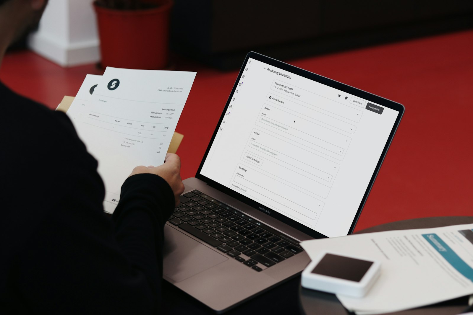 A paper invoice held next to a laptop showing a digital invoice being edited
